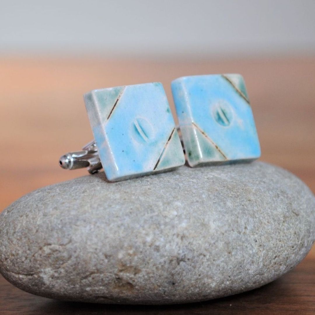 Two light blue ceramic square cufflinks placed on a pebble