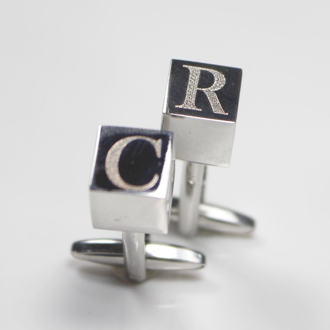 A pair of stainless stell cufflinks that are cube shaped and engraved with initials