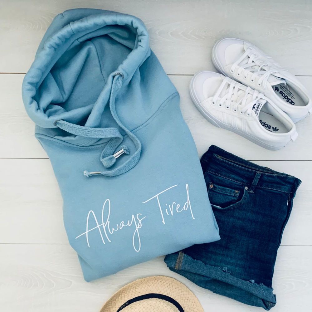two white sneakers, denim shorts, a hat and a plain light blue hoodie featuring the text "Always tired"