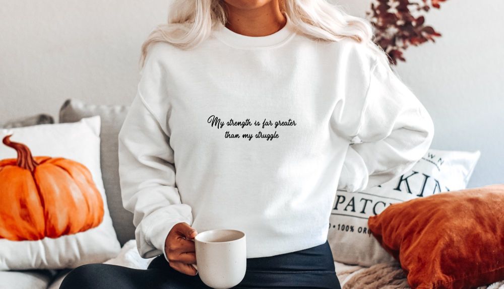 A plain white sweatshirt featuring the text "My strength is far greater than my struggle"