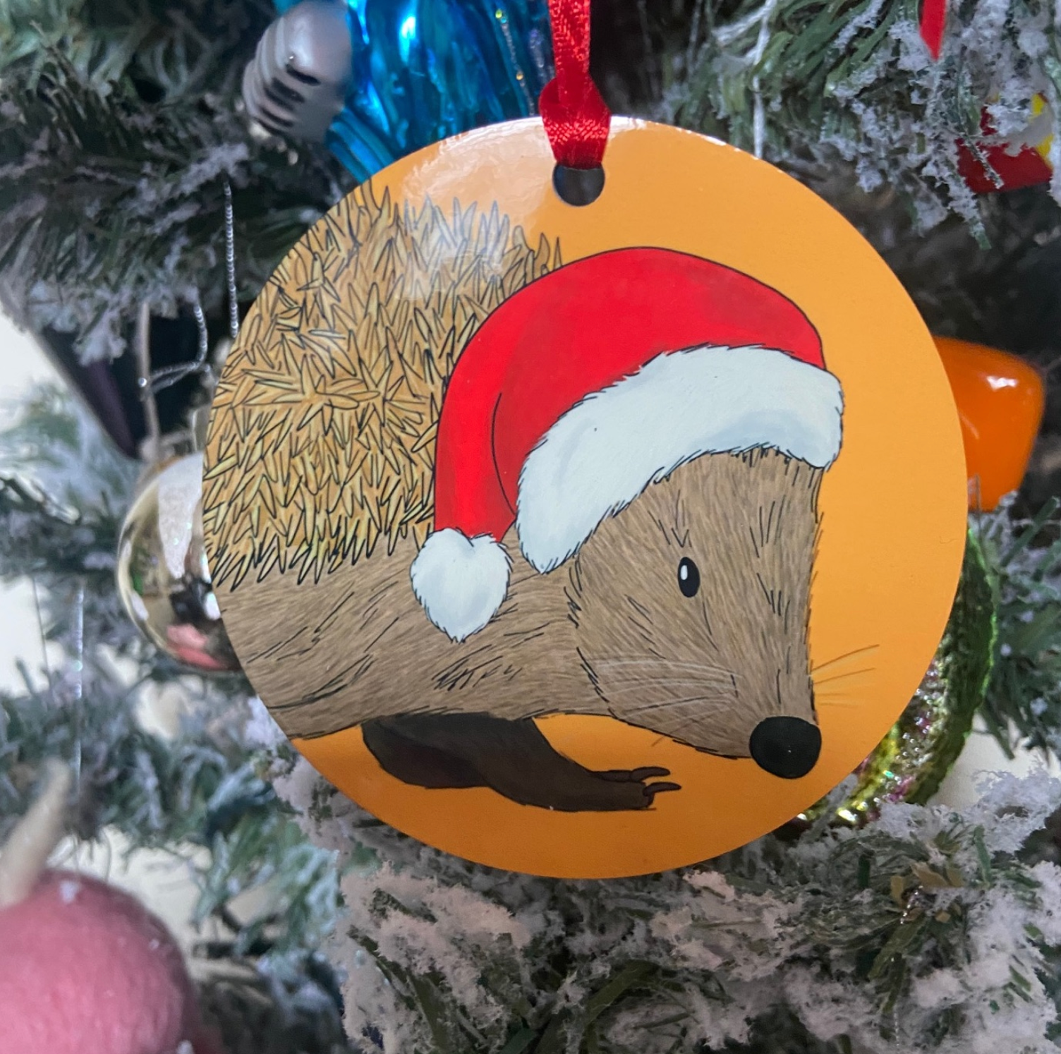 A christmas tree decoration featuring a handdrawn image of a hedgehog wearing a Christmas hat