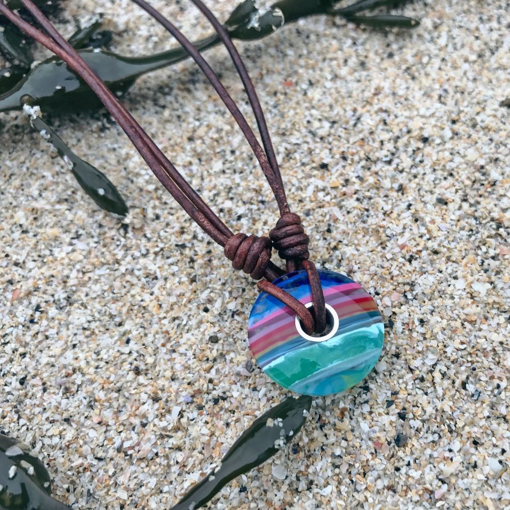 A multicoloured, polished circle pendant made from surfite on a dark brown leather necklace