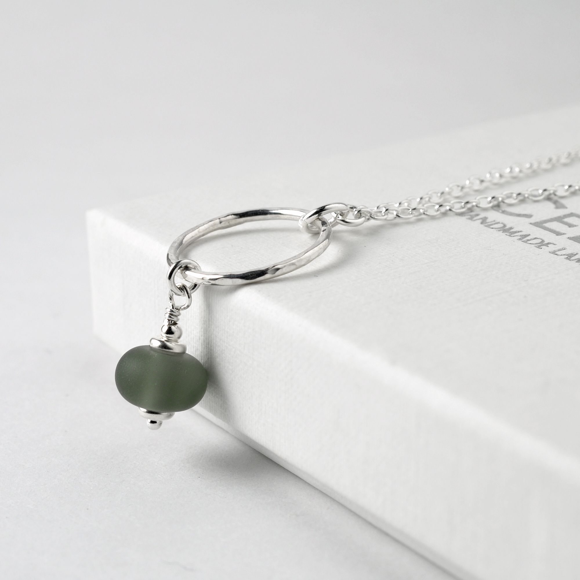 A slate grey tumbled glass pendant handing from a silver hoop on a necklace