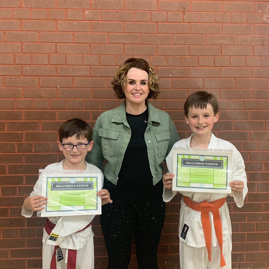 Two young boys in karate suits holding attendance certificates while smiling with their mother between them 