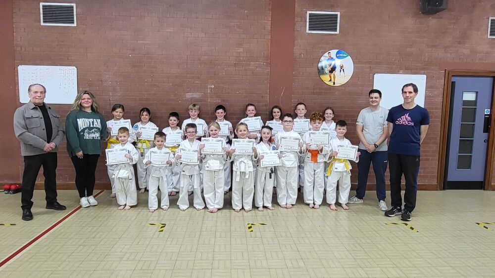 18 junior karateka standing against a wall smiling whilst holding certificates awarded for passing their belt examination. Theyu are flanked by two of their club instructors on each side