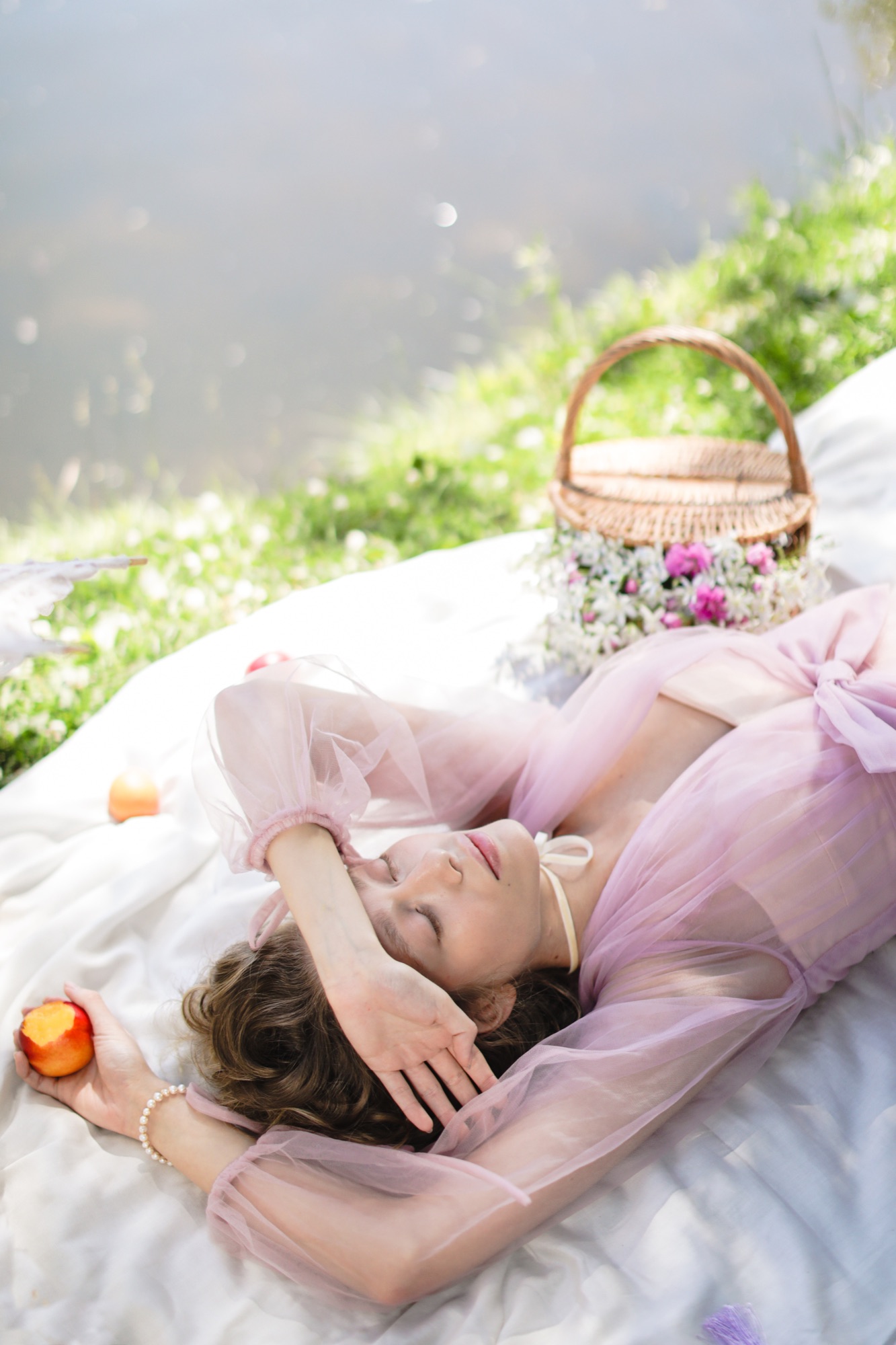 A young female wearing a pink and white dress. She is lying down carefree, on a pick-nick setting, holding in her hand a half-eaten peach. Her eyes are closed while enjoying the sunny weather, next to her is a beautiful pick-nick basket decorated with pink and white flowers.