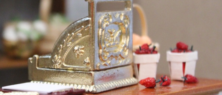 A miniature scene with a gold traditional till on a shop desk with strawberries on the side