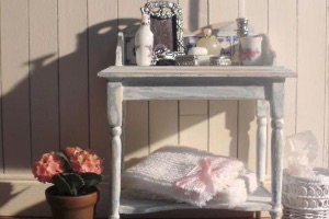 A dollshouse sized sideboard with miniature accessories and a plant, there are 12th scale towels underneath