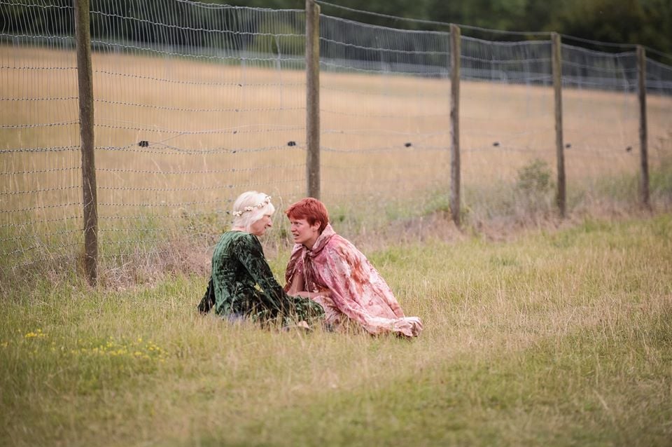 Outdoors on a grassy field, with a wire fince in the background. Two women are sitting on the grass. One is wearing a green dress, the other what looks like a repspattered cape. They look at eachoter intensly, while the women in a red clock is speaking 