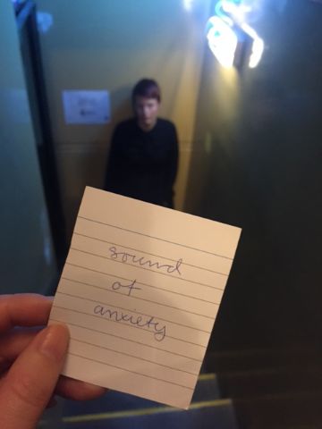Looking down a staricase, you can see a person dressed in black, out of focus. In the forground is a hand, holding a note whcih reads 'sound of anxiety' 