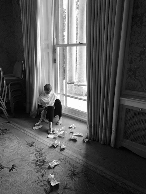 Artist sitting on the floor next to a llarge window. She is dressed in  white nightshirt, black leggings and barefoot. She is wearing a barely visible gag over her mouhth. Notepaper is scattered across the floor 