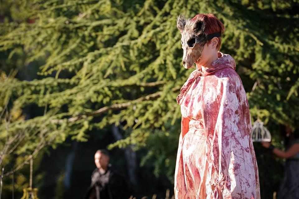 Artist wearing a wolf head mask and a cape stained with red spatters, standing outdoors