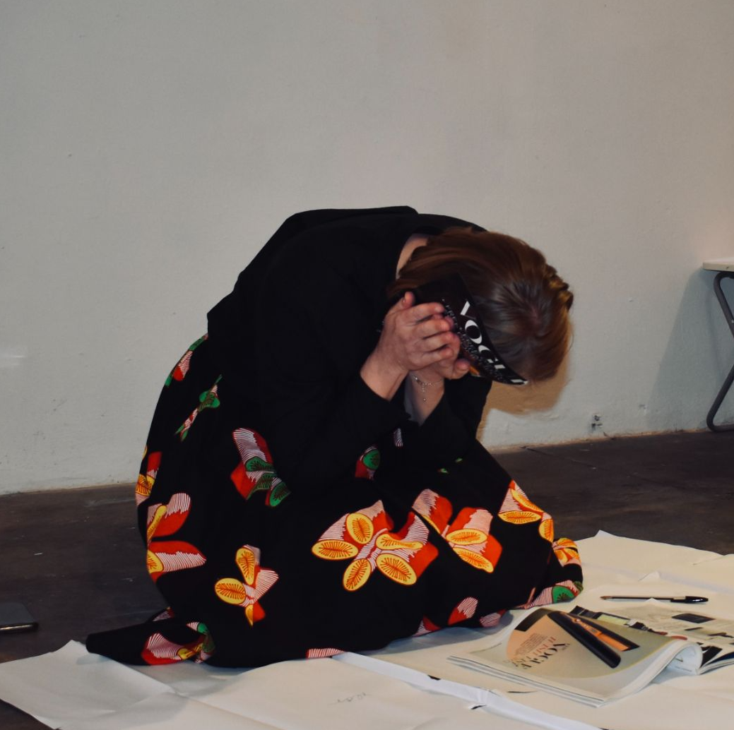 Female artist kneeling on the floor, holding a magazine page pressed to her ear 