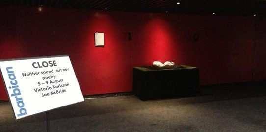 A red wall with a table in front. On the table is a pillow, resting on wat looks like soil and grass. 