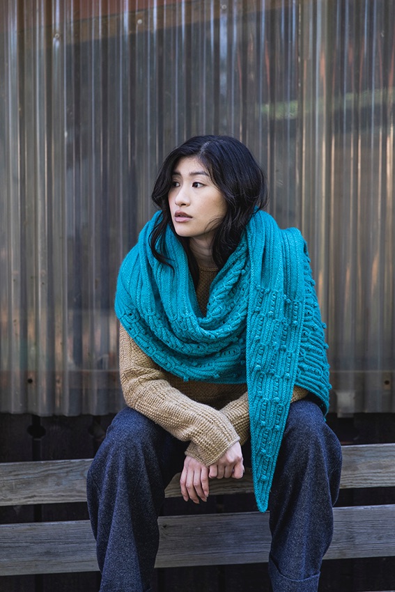 Young Asian woman  sitting in front of corrugated wall wearing large turquoise coloured Teira shawl, beige top and navy trousers