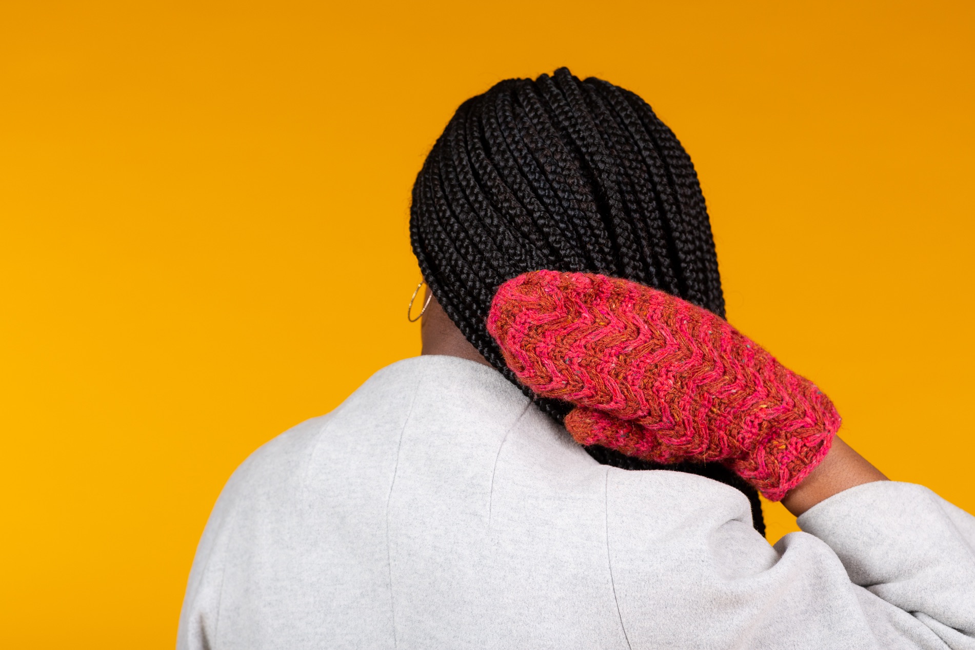 Back view of young african girl's braided hair with  right hand wearing brightly coloured Toasty Cosy knitted mittel, white coat against bright yellow background. 