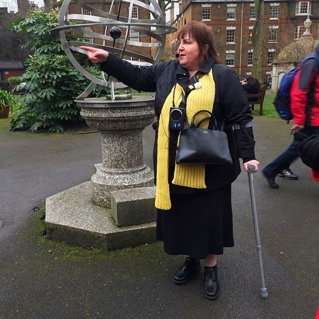 Photo of Tina leading a gardens walk for Baker Street Quarter Partnership. Tina is wearing a black coat and skirt with a yellow scarf. She stands holding her crutch and pointing to an object which is out of view of the photo. In the background there is a medium size metal sundial on a marble plynth.