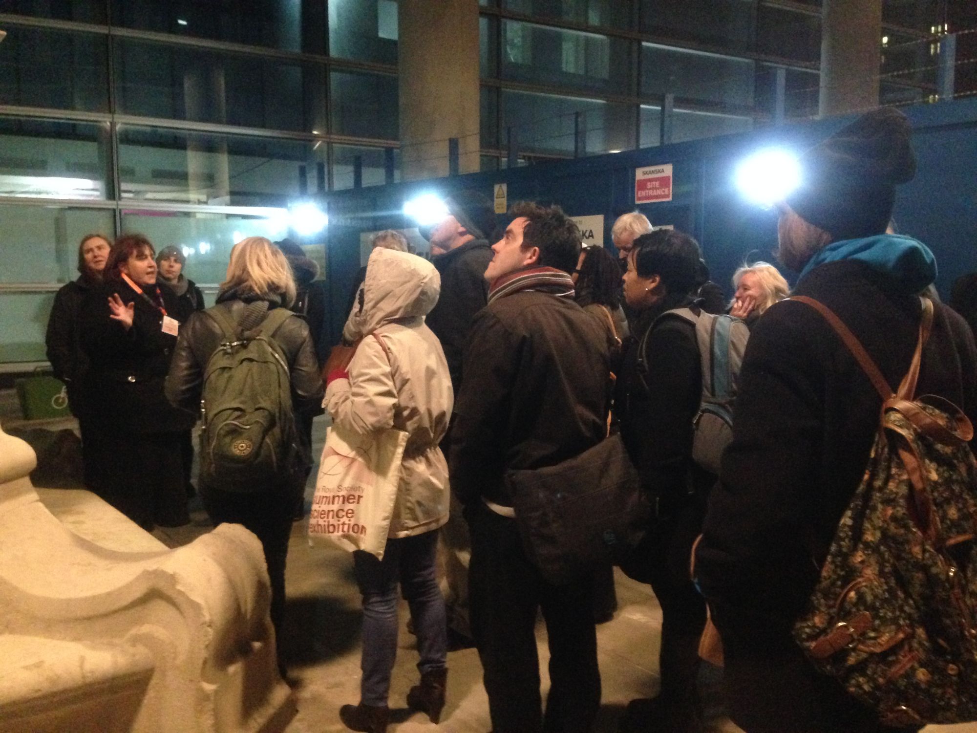 Photo of Tina leading a medical history tour  for Centre of the Cell.  The small group is standing at the base of a statue in the grounds of the London Hospital, Whitechapel. In the background is some temporary boarding, as there was building work taking place . The tour took place in the evening, so it is dark and the street lamps are lit.