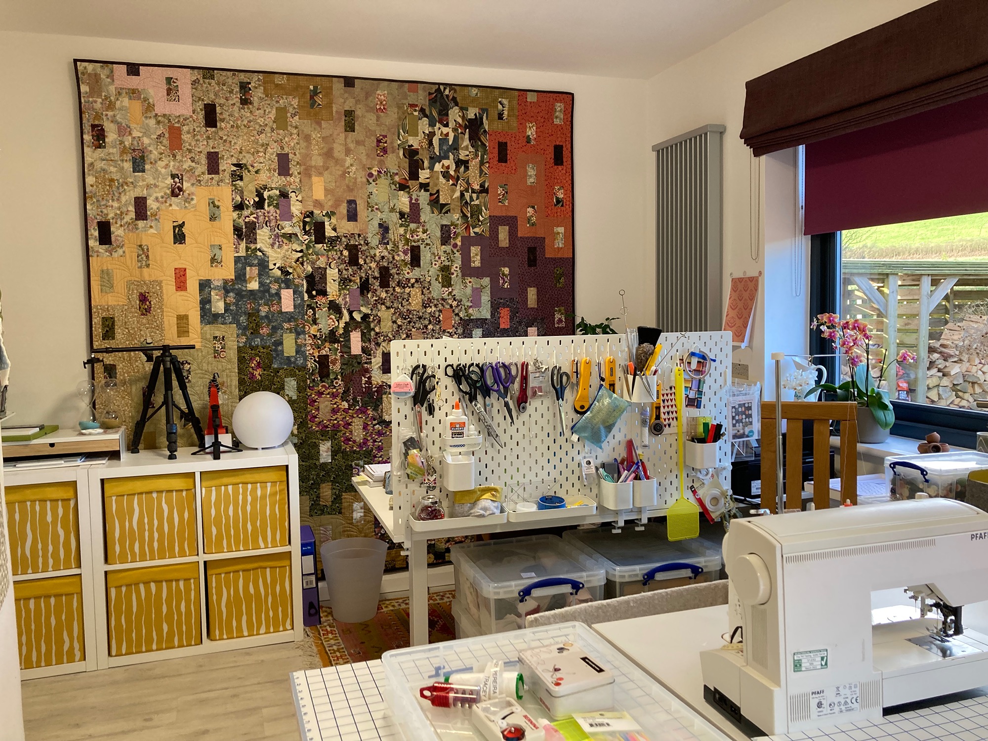 Image of a purple and gold quilt on a wall with a desk and tools in front of it