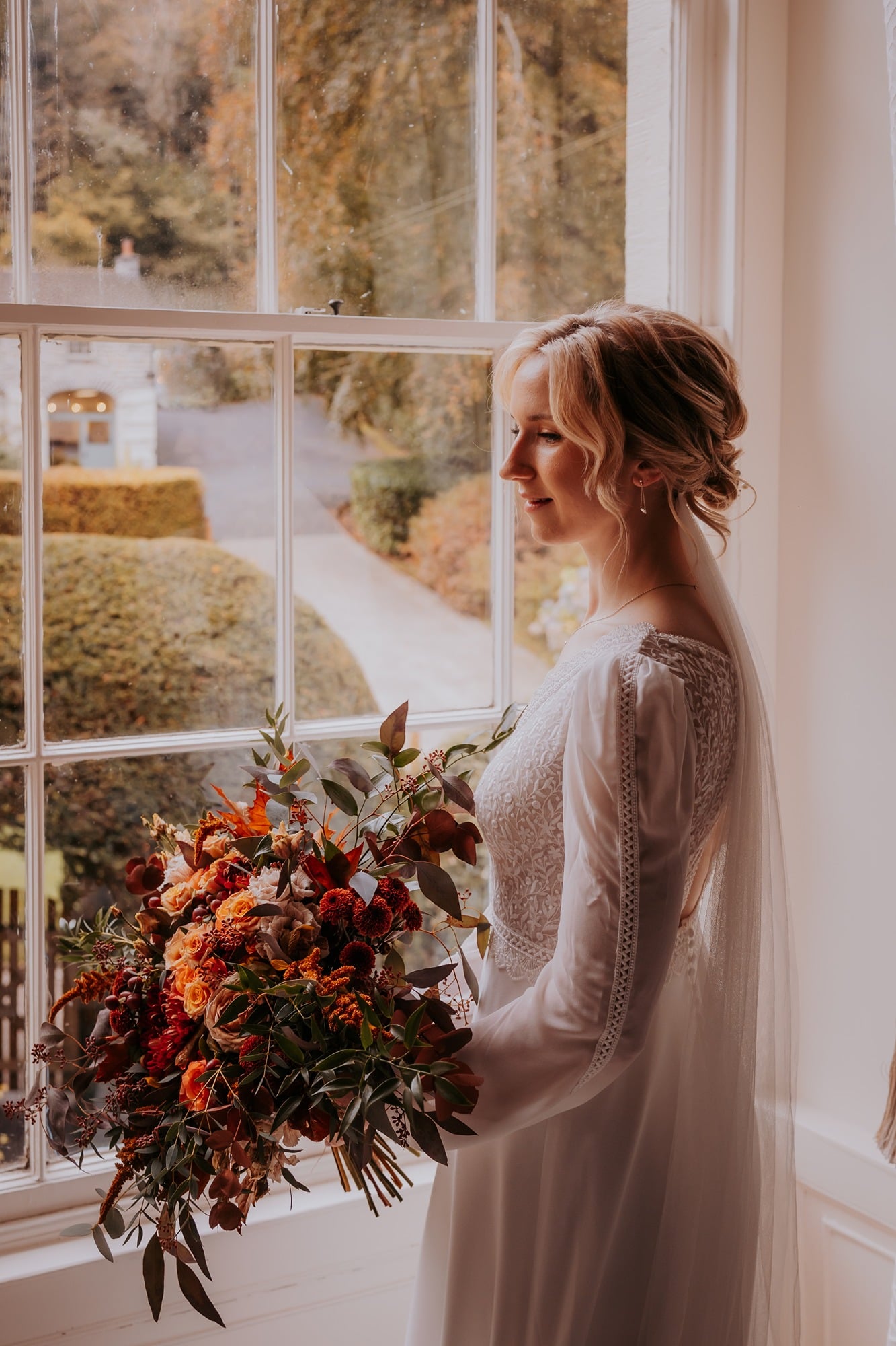Bride with Autumnal bouquet looking out of bridal suite window, Town Head Estate, Windermere
