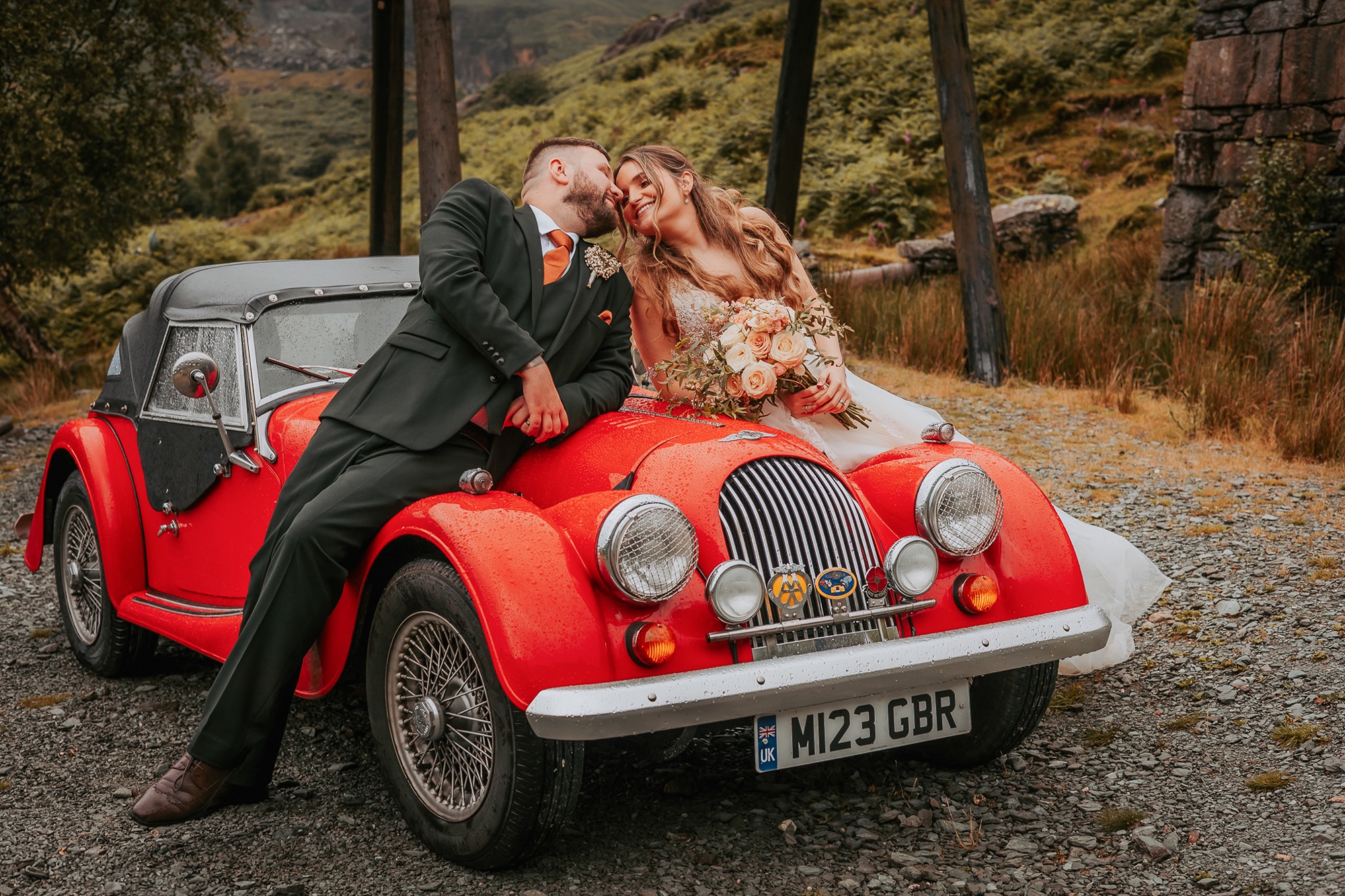 Bride and Groom embrace in a kiss over the bonnet of a vintage red car