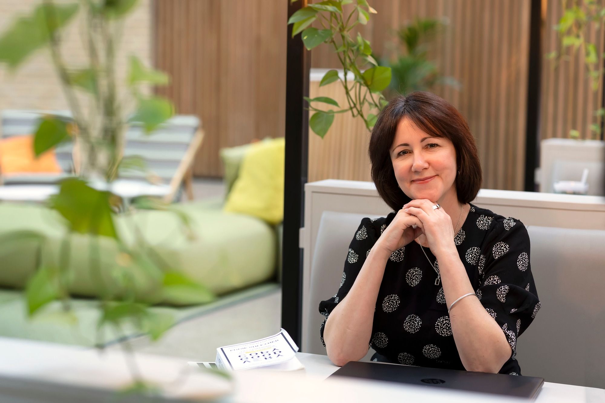 A professional freelance copywriter sits at a desk with a laptop. She is looking at the camera and smiling