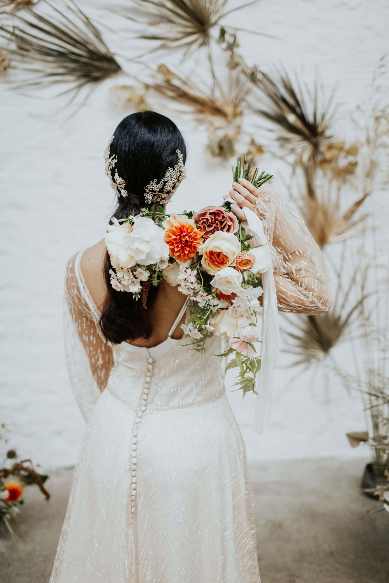 A model stands with her back to us. She has a bouquet of ivory and coral flowers over her shoulder, she is wearing a Rachel Burgess Bridal Boutique wedding gown of gold glittery lace and is wearing a delicately beaded bridal headpiece made by Clare Lloyd Accessories