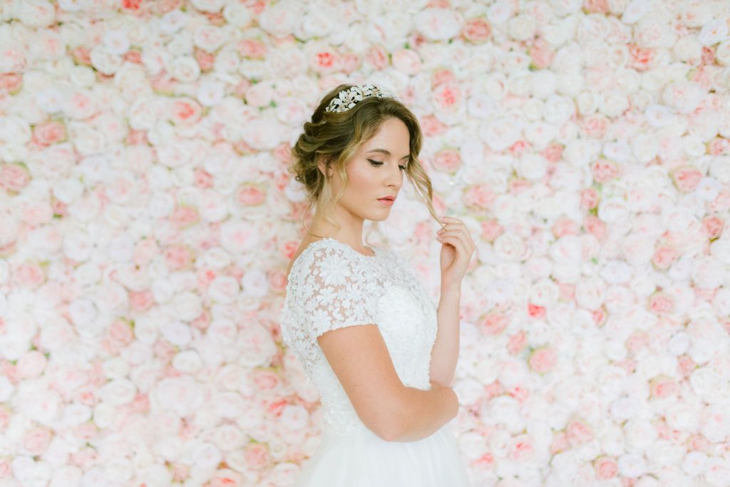 A bridal model stands infront of a floral wall of pink roses with a lace wedding dress and a white leaf qedding crown