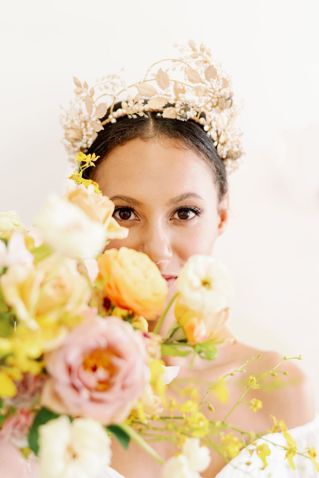 A model wears a statement pale gold and pearl wedding crown headdress and holds a bouquet of pastel coloured flowers
