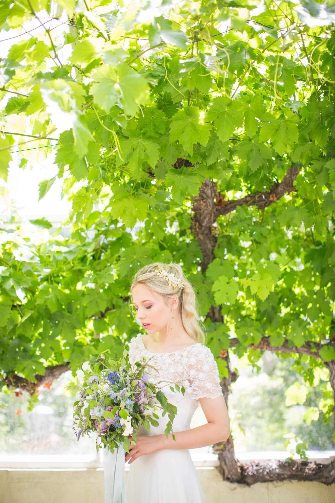 A bridal model holds a summer flower bouquet and stands under a fig tree in an English country garden 