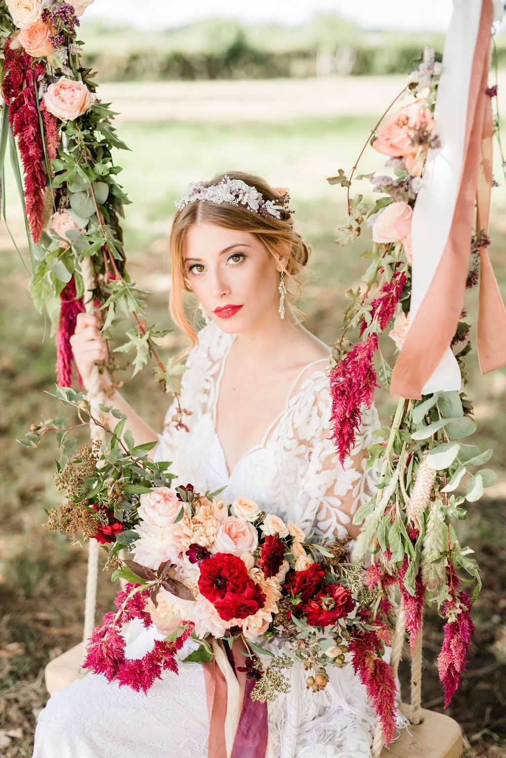 A bridal model sits on a swing suspended with flowers and foliage and holds a bouquet of red and coral flowers