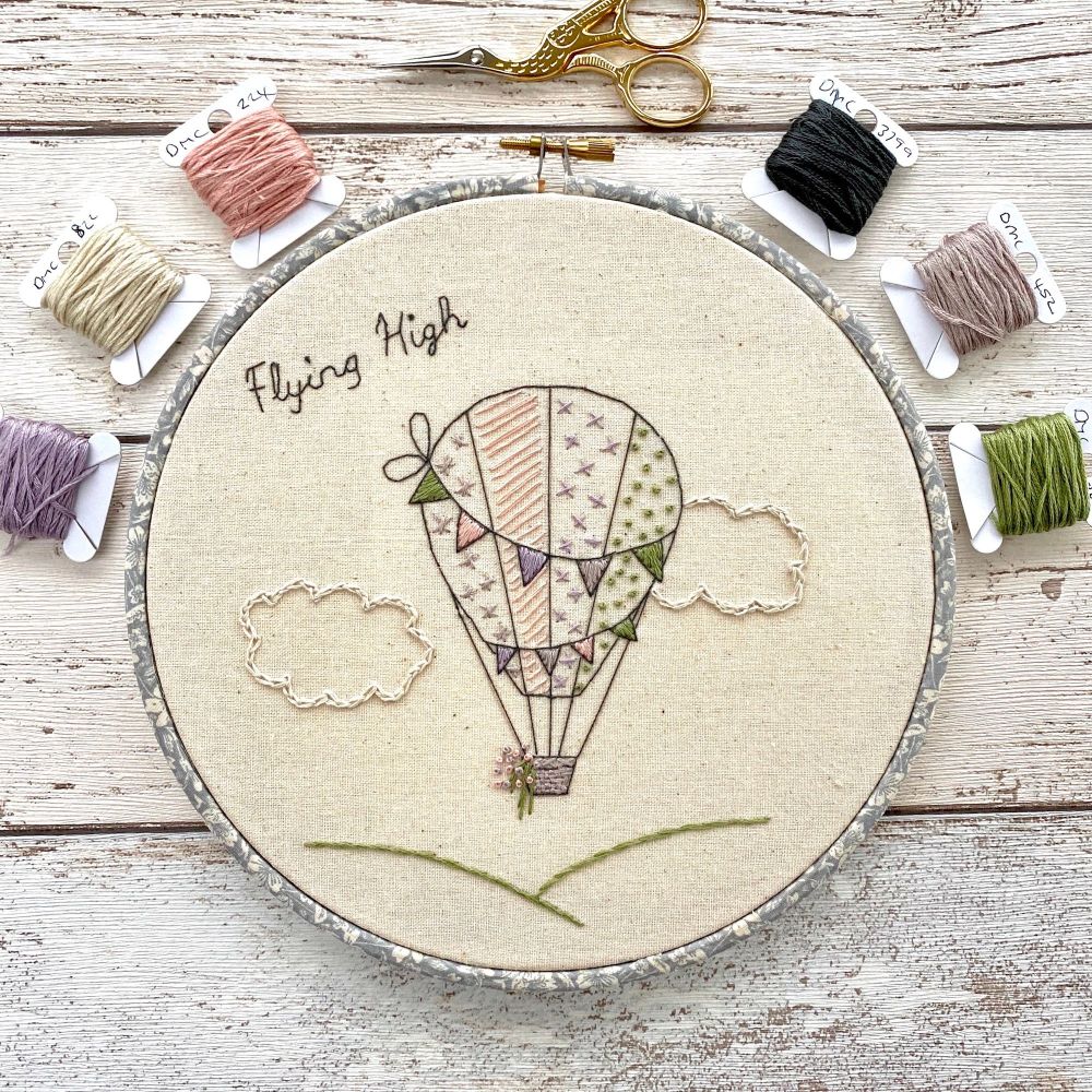 A photo of an embroidery hoop of a hot air balloon and the words flying high.  A selection of threads and a pair of scissors are laid around the hoop on a wooden background