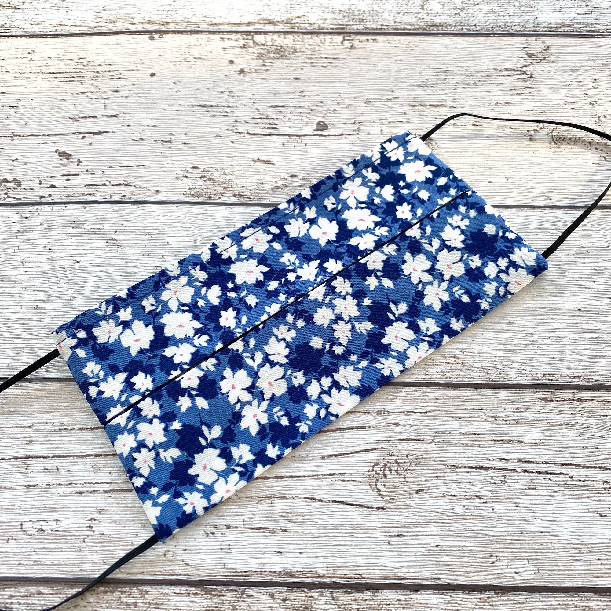 A mid blue coloured face mask with a pattern of  white and navy flowers.  Black elastic at each end lying on a wooden surface.