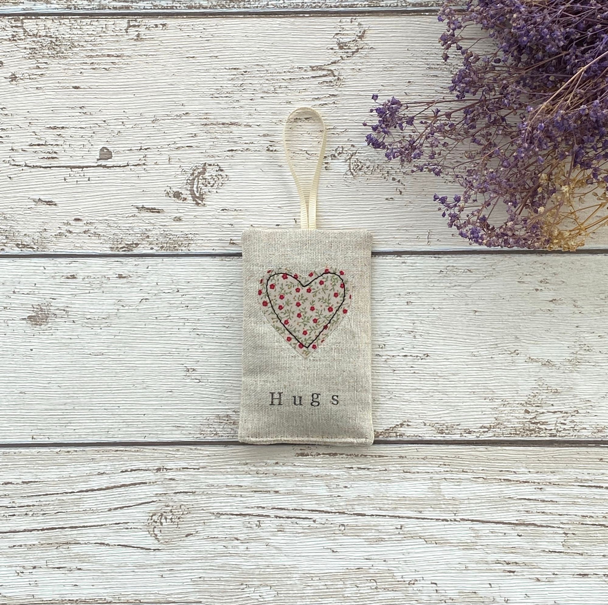 A rectangle linen bag with a fabric heart stitched to the front. The heart has little red flowers on. A cream ribbon is fixed at the top.  SHown on a wooden surface with purple dried flowers.