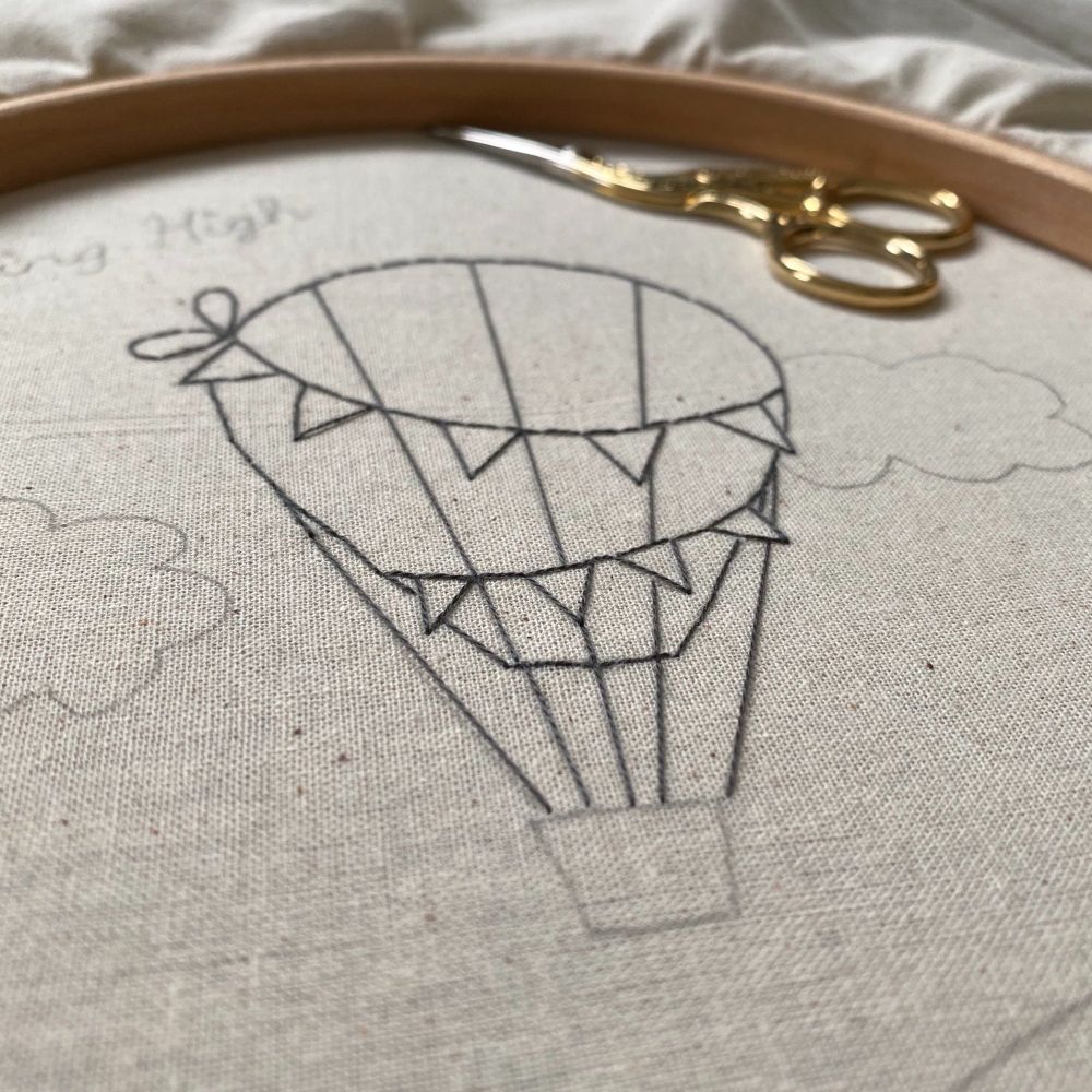 A work in progress embroidery hoop showing outline stitching of a hot air balloon picture in black thread, a small pair of scissors are lying towards the edge of the hoop.