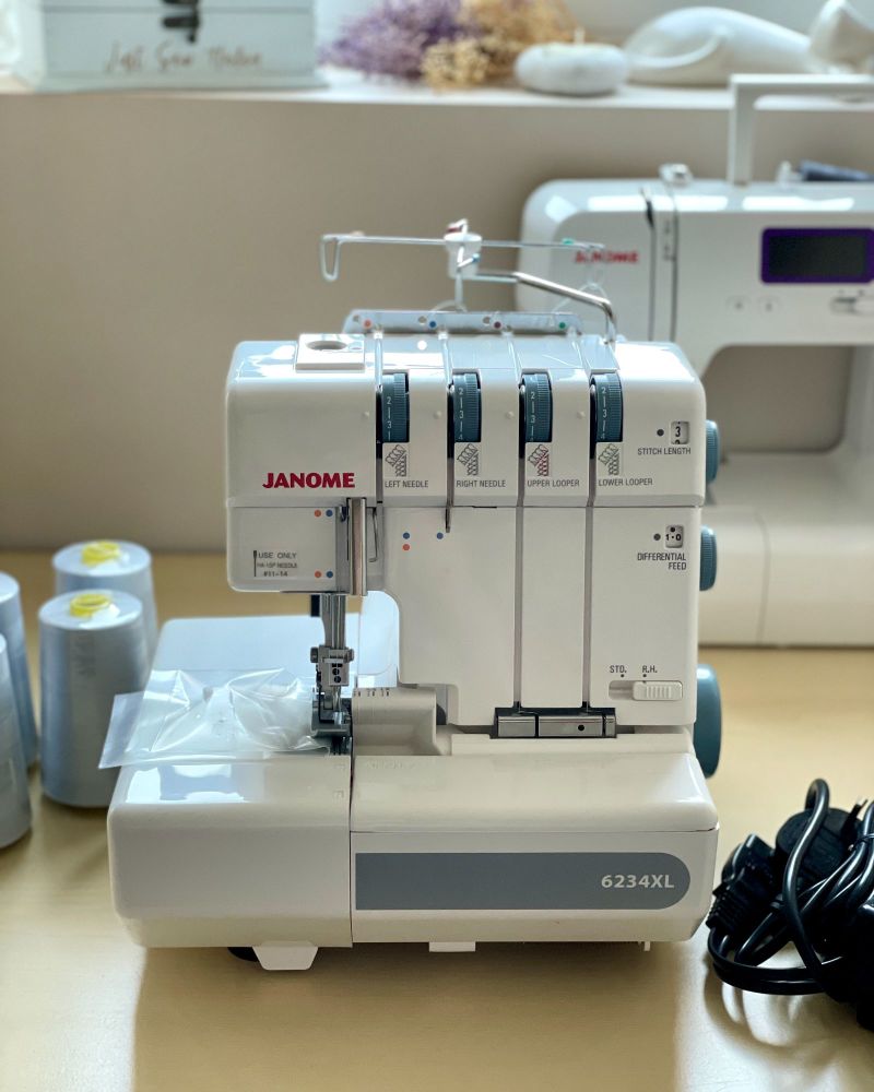 A Janome Overlocker 6234XL machine on a desk with spools of cotton. There is another Janome sewing machine behind it, below a window sill.