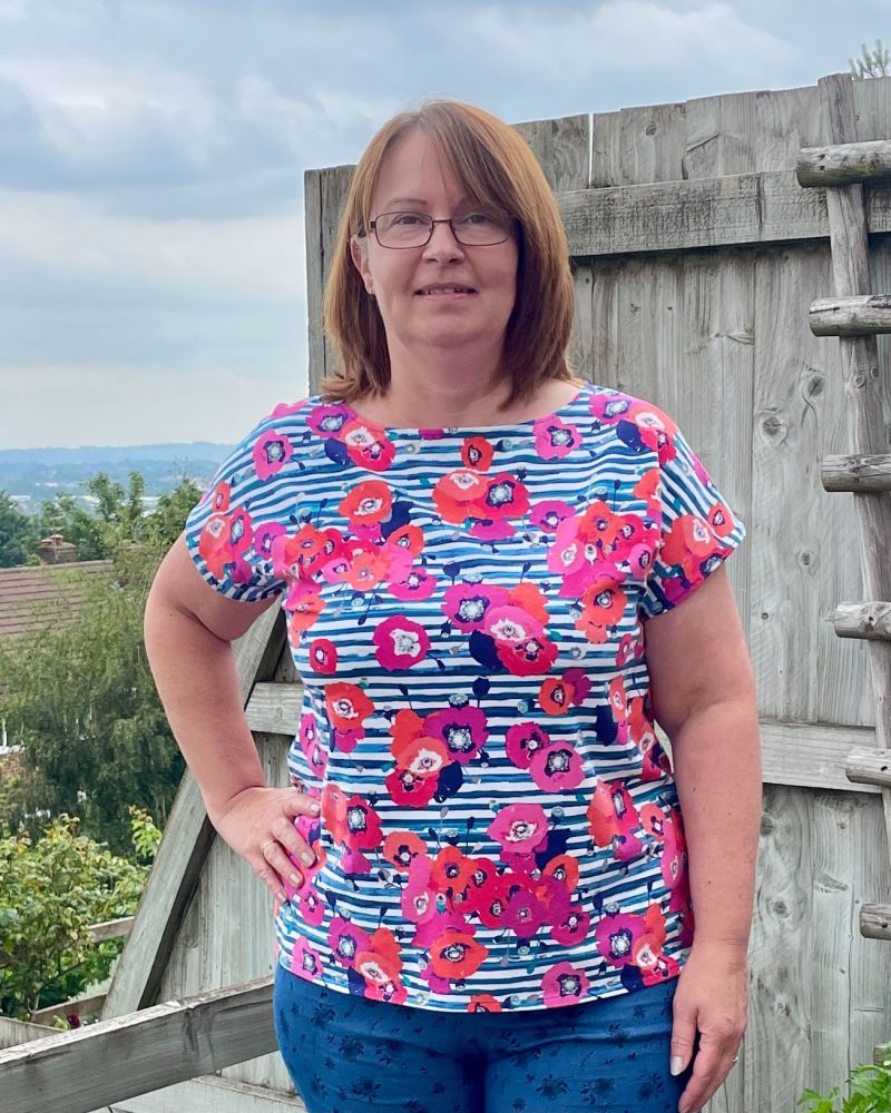 A lady with brown hair and glasses is wearing a blue stripe and floral tee shirt, standing in front of a garden fence.