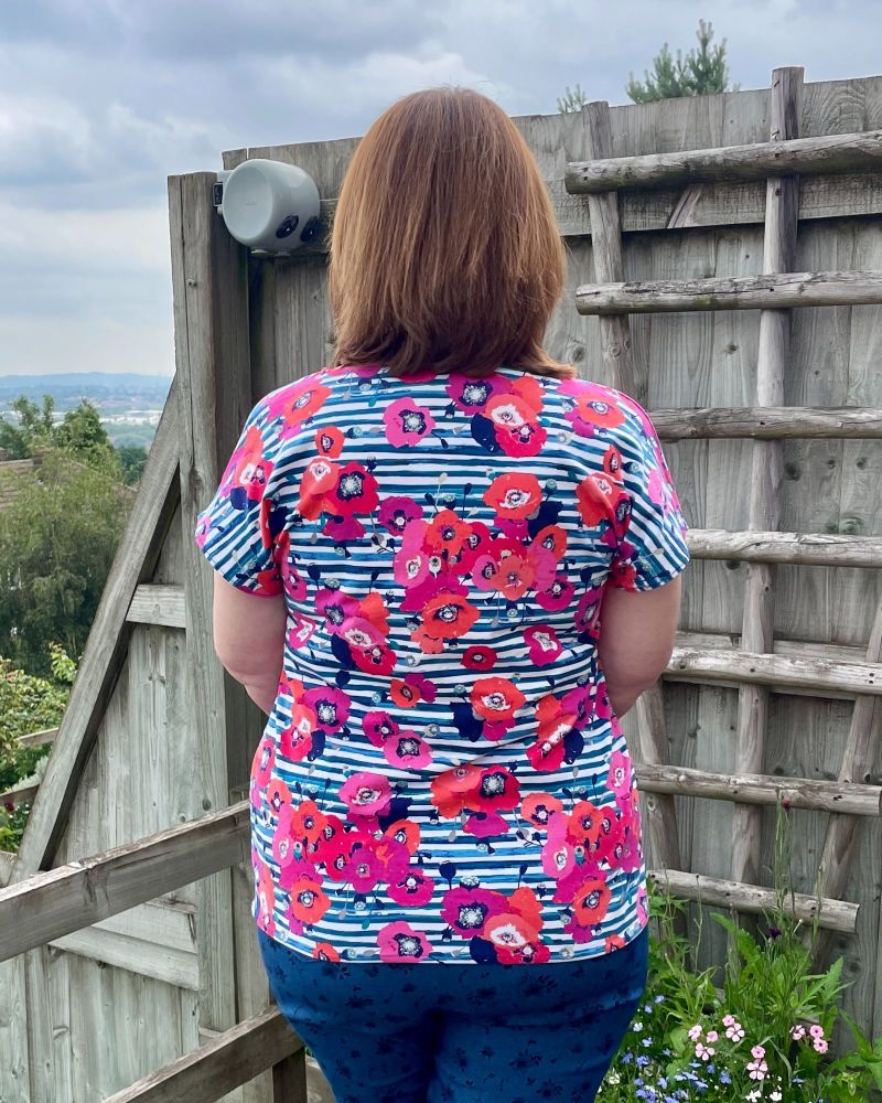 A lady with brown hair is wearing a blue stripe and floral tee shirt, standing in front of a garden fence.