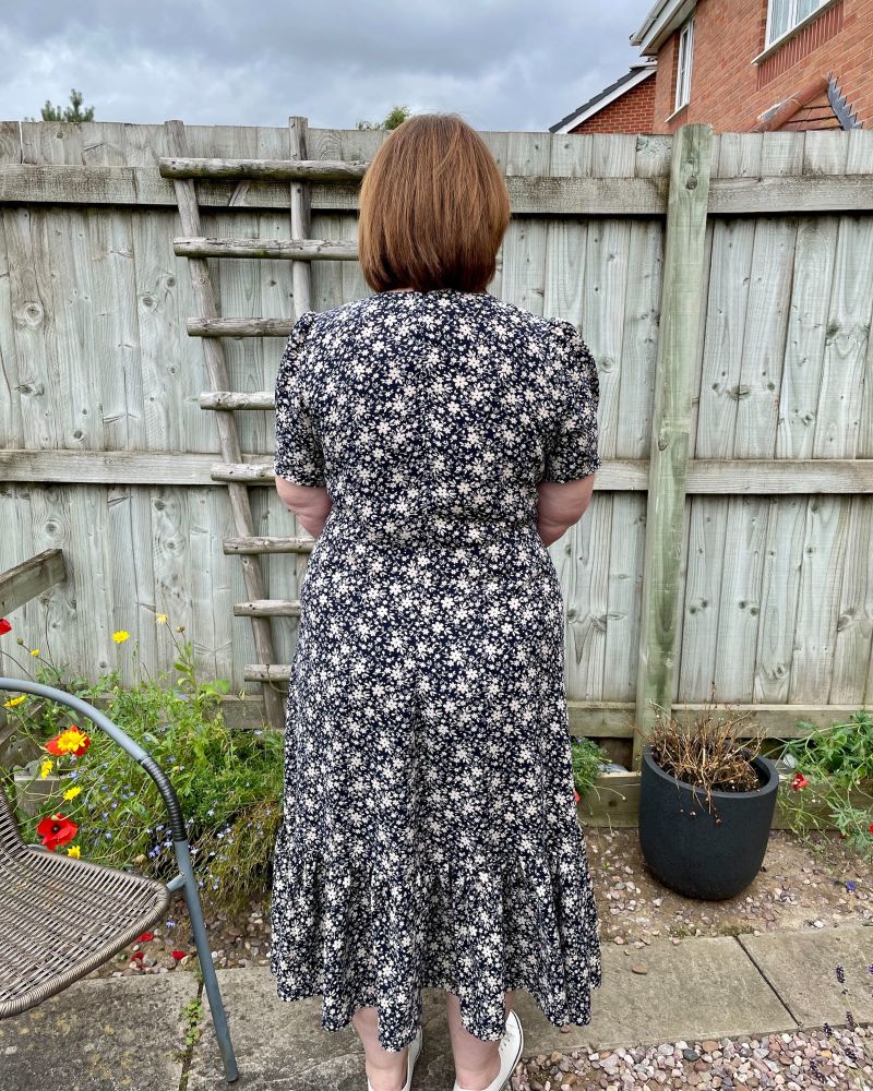  A lady wearing a navy floral dress made using sewing pattern Simplicity 8875 showing the back view and standing in front of a garden fence.