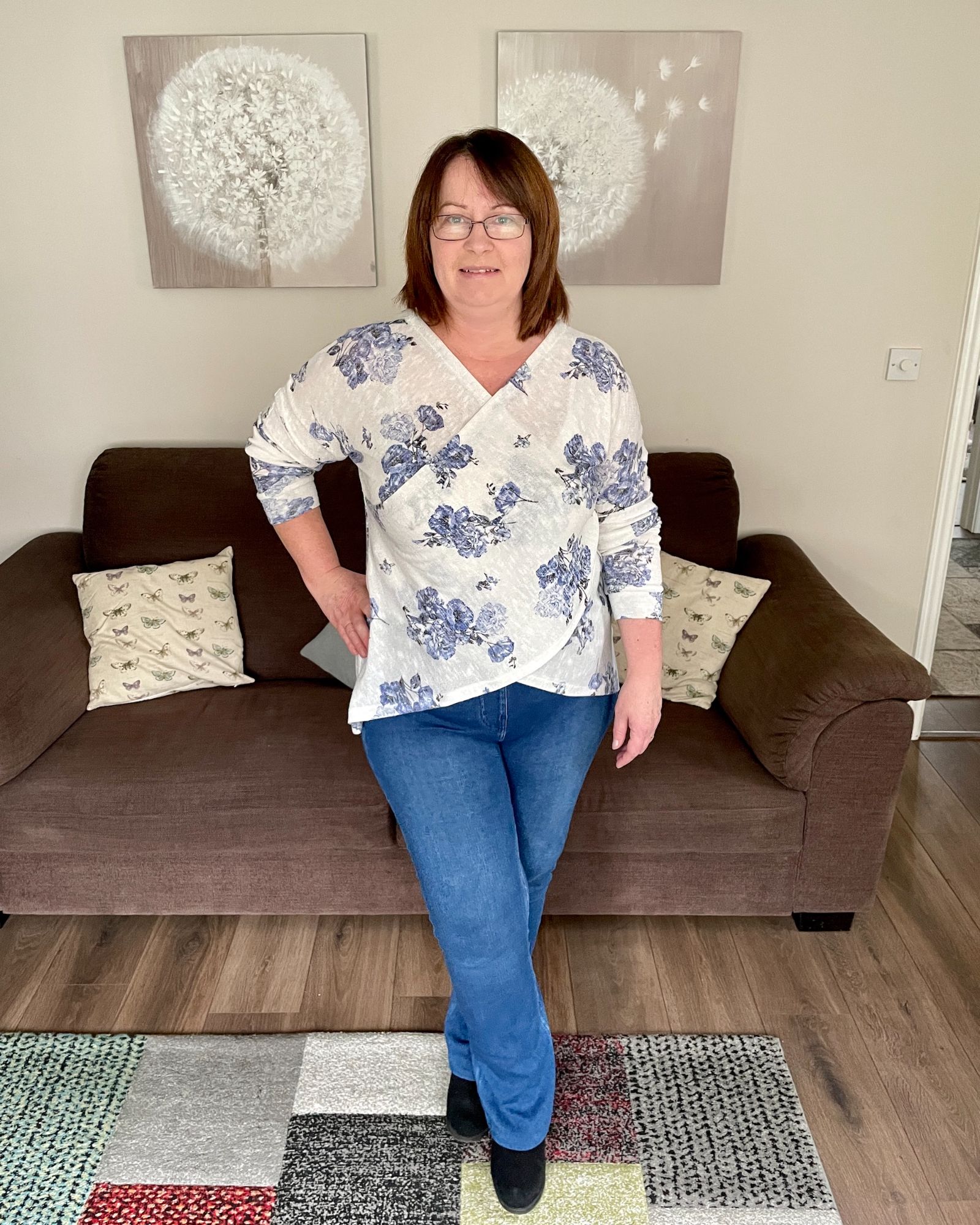 A lady standing in the living room wearing blue jeans and a blue floral and ivory cross over jumper