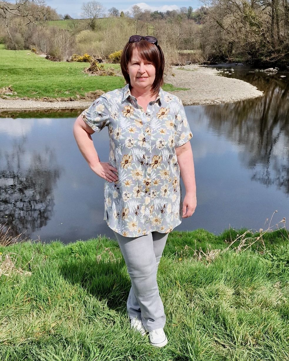 A lady standing in front of a river wearing a grey floral shirt with grey jeans.