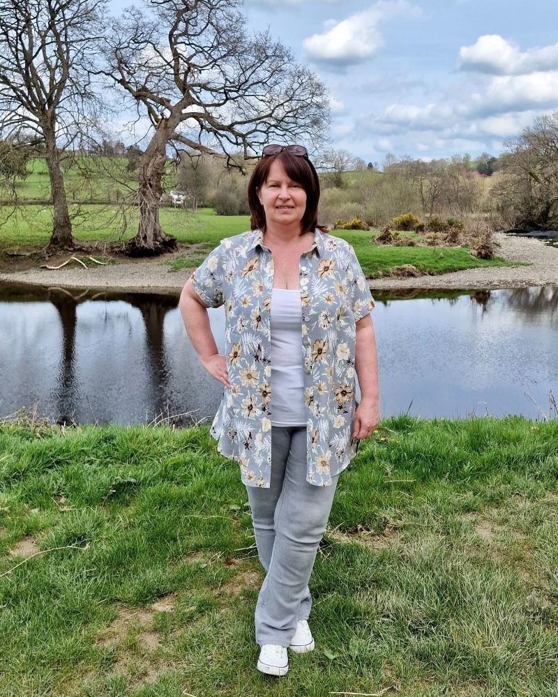 A lady standing in front of a river in the sunshine wearing a grey floral shirt and grey jeans