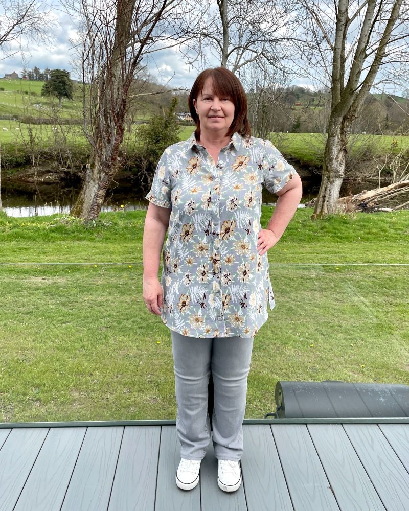 A lady standing in the countryside wearing a grey flowered shirt and grey jeans