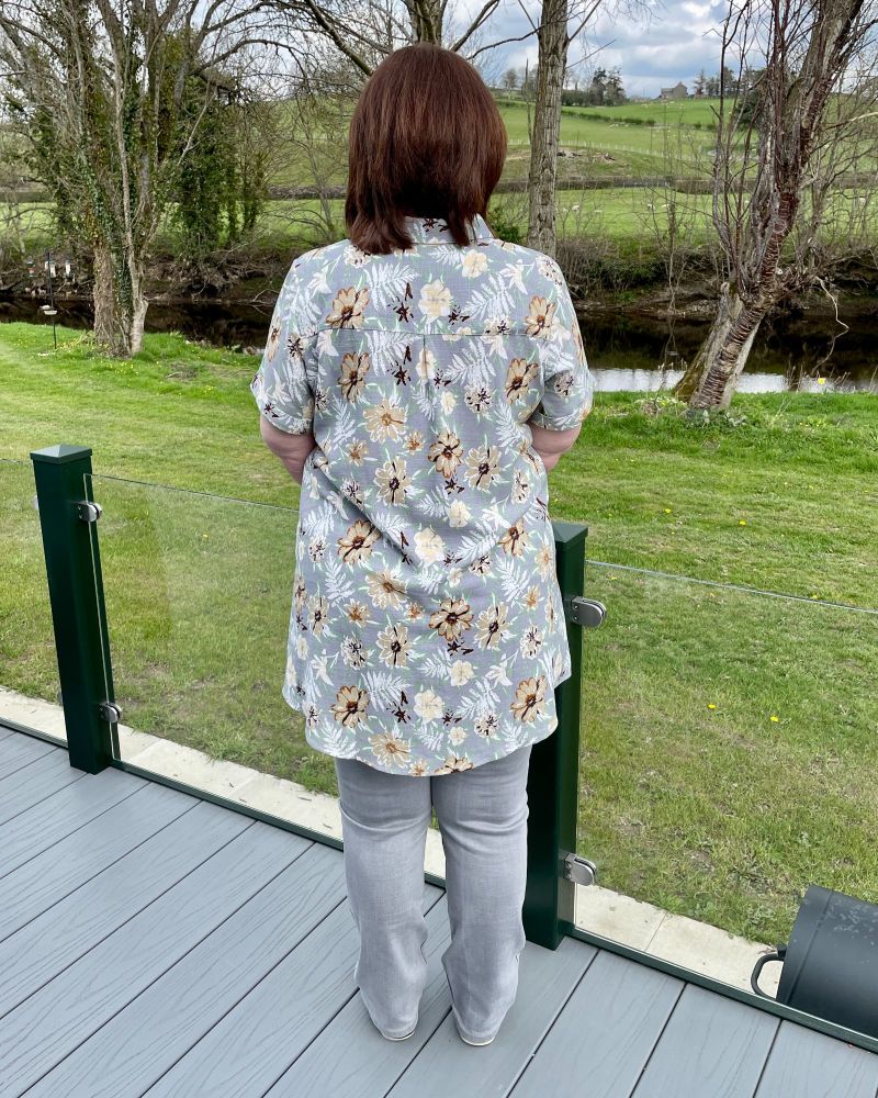 A lady standing with her back to the camera in the country side wearing a grey flowered shirt, grey jeans and white pumps