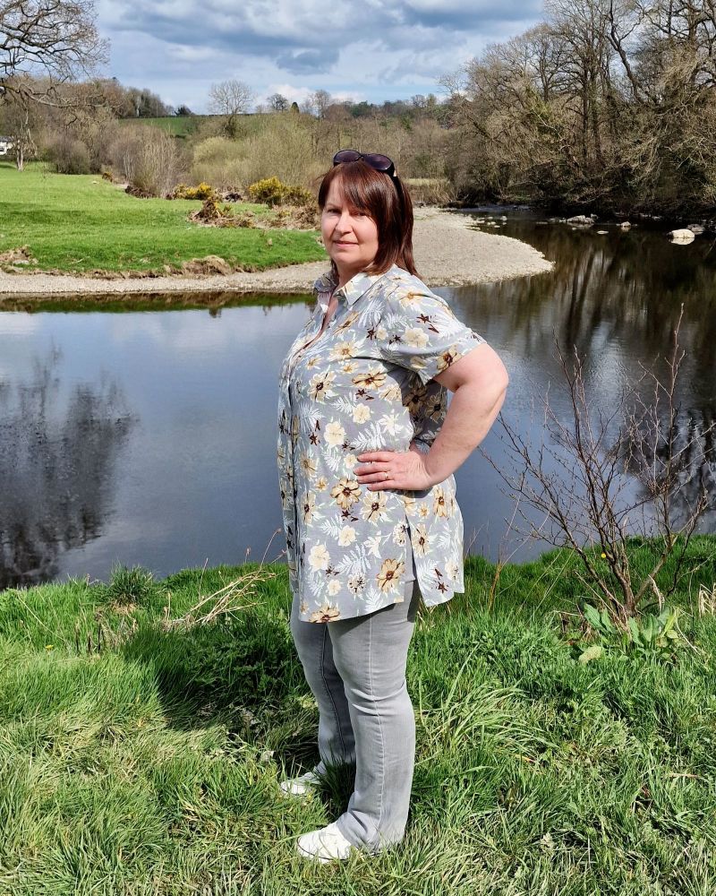 A lady standing in front of a river showing the side view of her grey flowered shirt