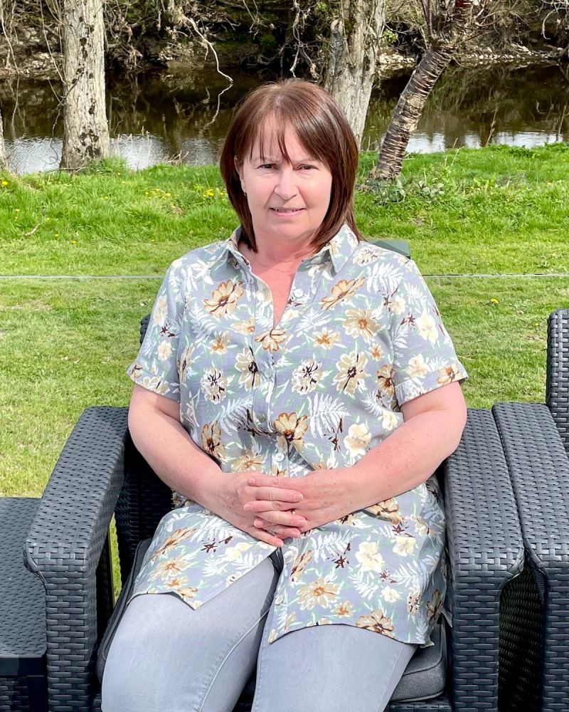 A lady seated by a river wearing a grey floral shirt and grey jeans