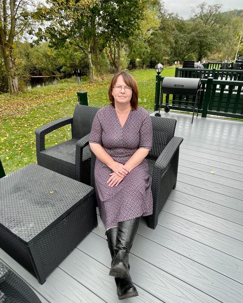 A lady sitting on garden furniture on decking wearing a black and pink Dress Shown seated