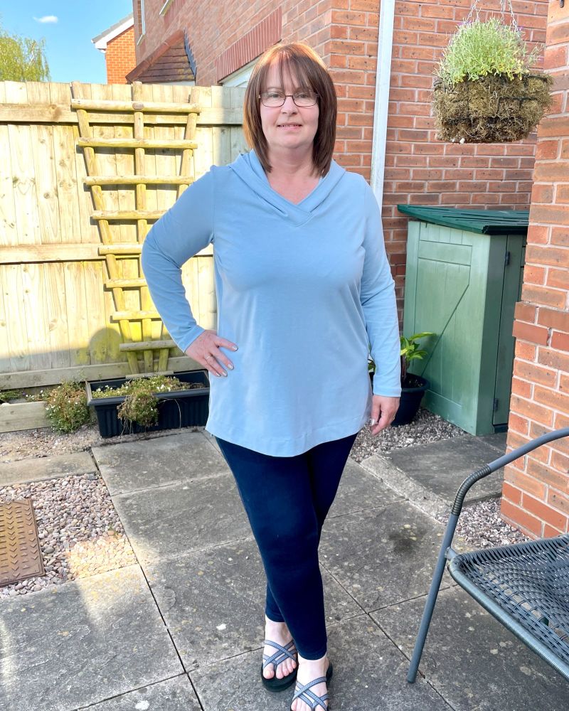 A lady standing in a garden in front of a fence wearing a light blue tunic top