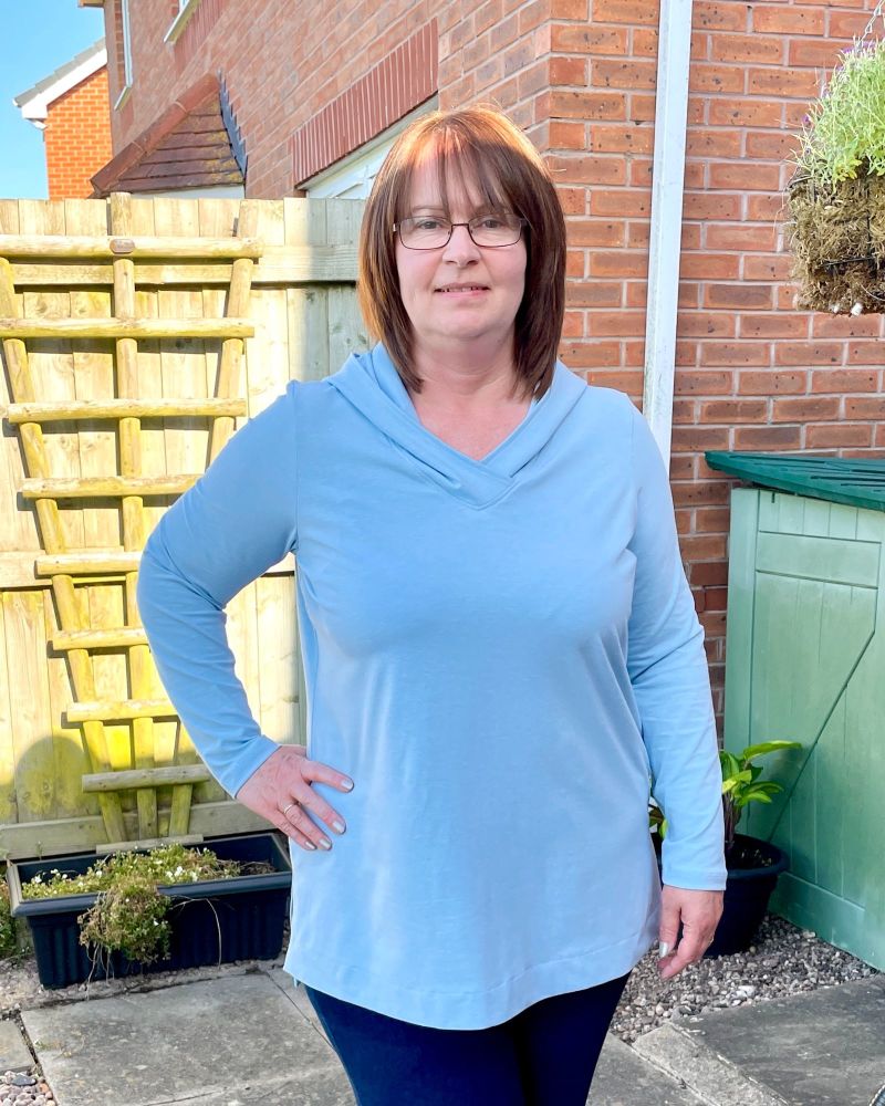 A lady standing in a garden in front of a fence wearing a light blue tunic top