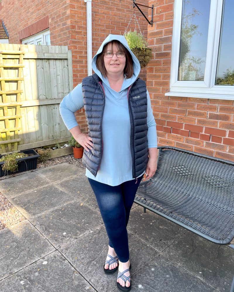 A lady standing in a garden in front of a fence wearing a light blue tunic top with the hood up and wearing a body warmer on top.